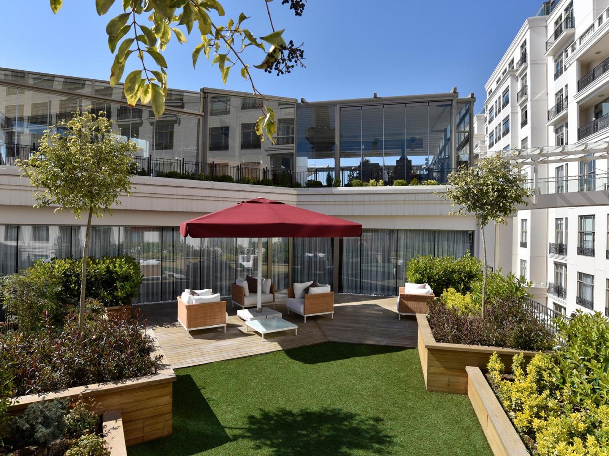 Cvk Park Bosphorus Hotel Istanbul Room photo The photo shows a stylish outdoor area set in a modern building. It features a well-manicured lawn surrounded by decorative wooden planters filled with plants. In the center, there is a large red umbrella providing shade over a seating area, likely d