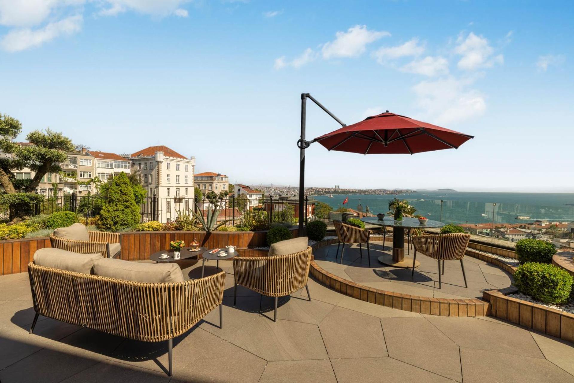 Cvk Park Bosphorus Hotel Istanbul Exterior photo The photo shows a scenic outdoor terrace or patio area with comfortable seating arrangements. There are several woven chairs and a couple of tables, creating a cozy atmosphere for relaxation or socializing. A large red umbrella provides shade, and th