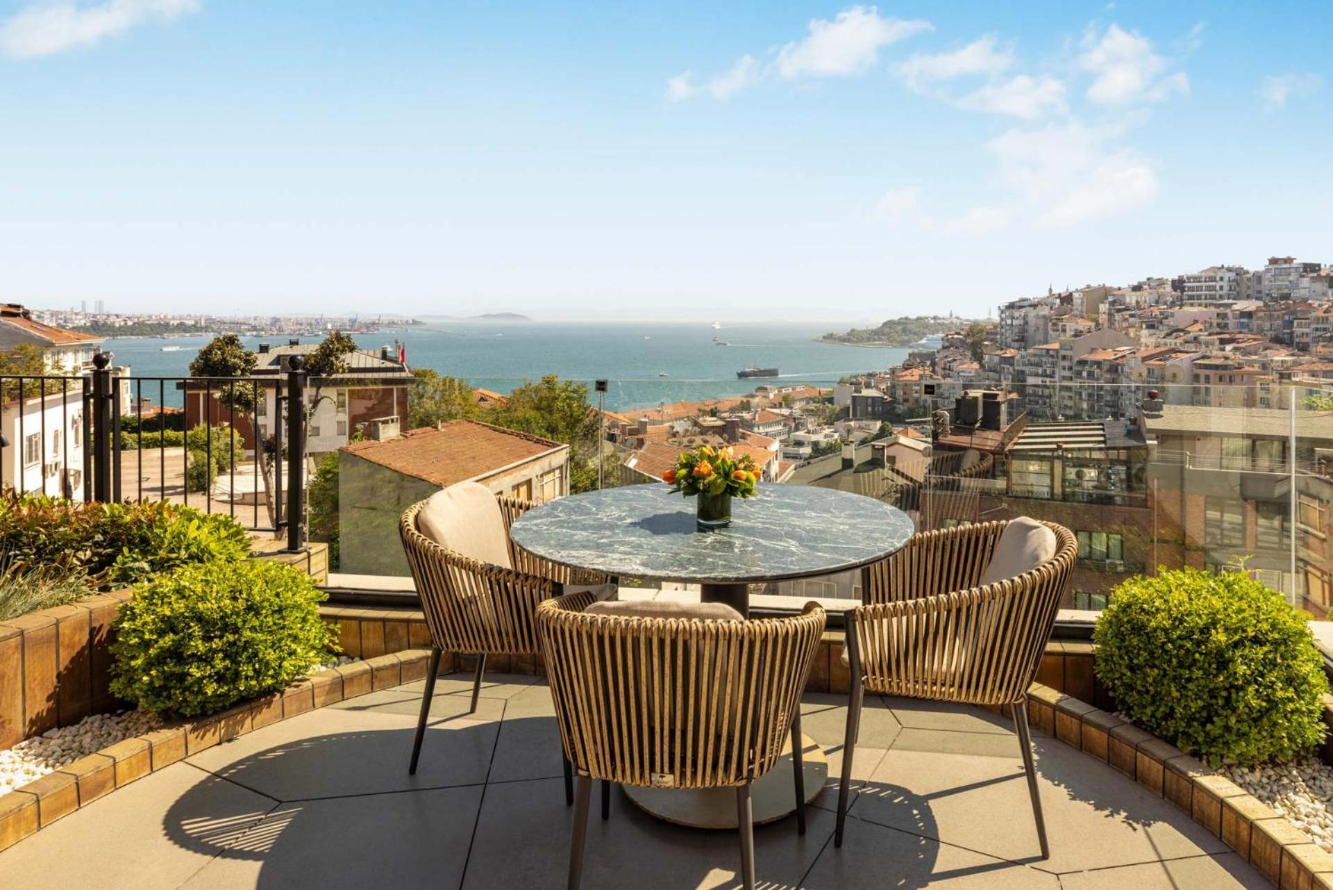 Cvk Park Bosphorus Hotel Istanbul Exterior photo The photo shows a terrace with a stunning view of a body of water and a cityscape. There is a round table made of a stone-like material, surrounded by four wicker chairs. A small potted plant with flowers is on the table. In the background, you can s