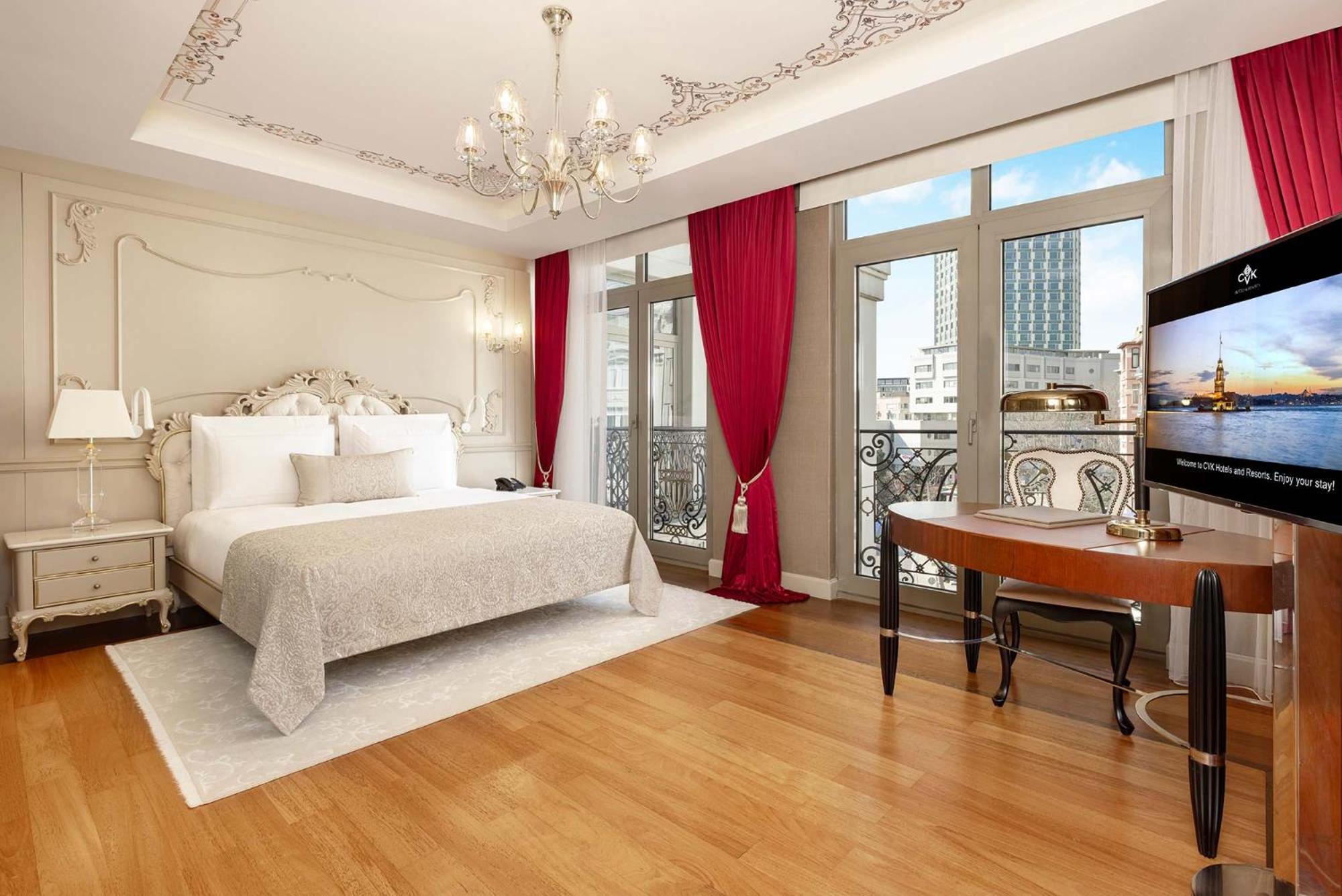 Cvk Park Bosphorus Hotel Istanbul Exterior photo The image shows a beautifully decorated hotel room. It features a large bed with a light gray cover and white pillows. The walls are adorned with elegant moldings and a stylish chandelier hangs from the ceiling. There are large windows with red curta