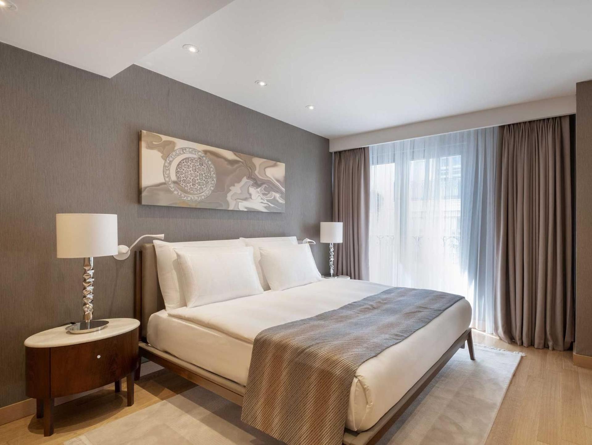 Cvk Park Bosphorus Hotel Istanbul Exterior photo The photo shows a modern hotel room featuring a large bed with white bedding and a light grey throw blanket. On each side of the bed, there are stylish bedside tables with lamps. The walls are painted in a soft grey tone, and there is a piece of abst