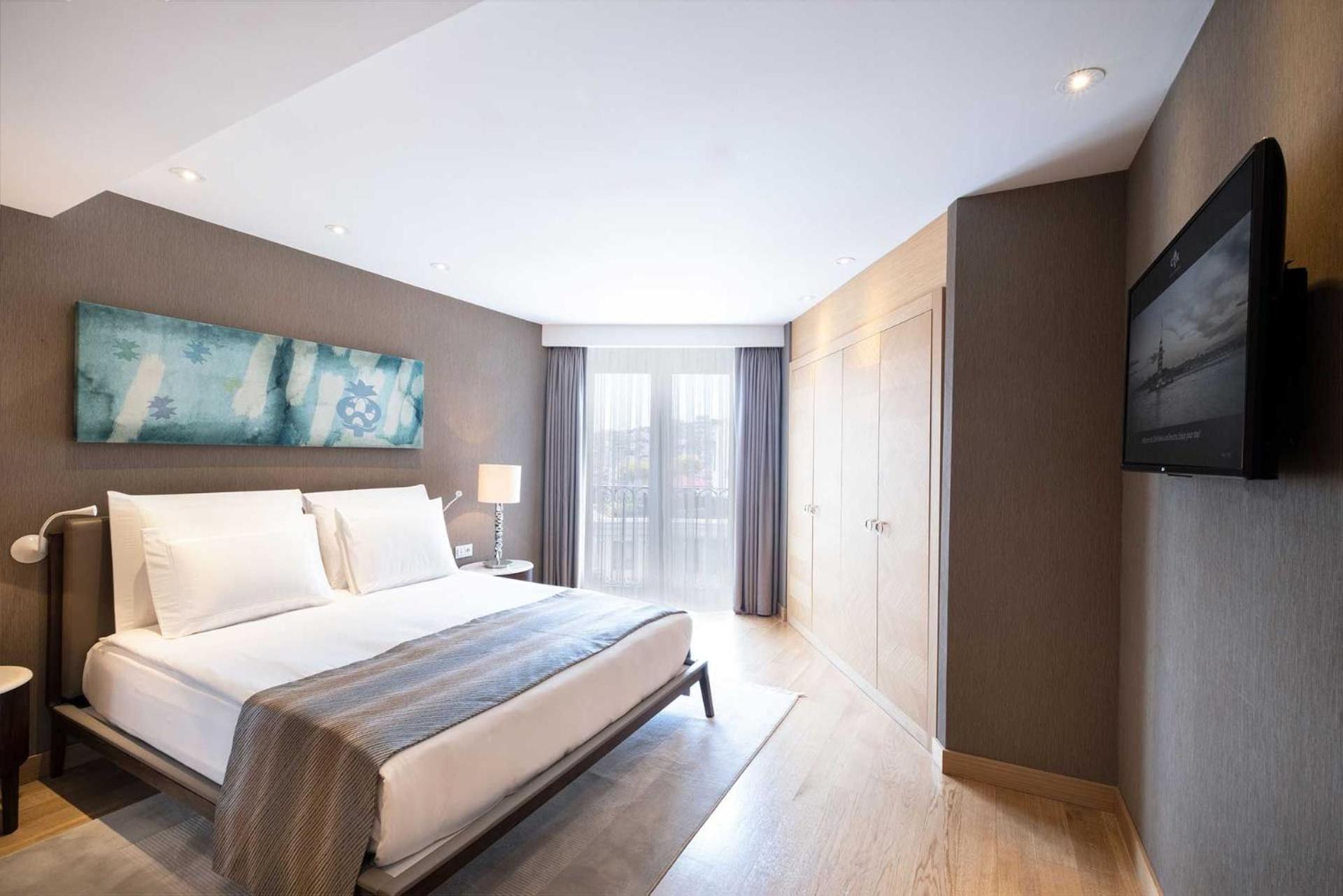 Cvk Park Bosphorus Hotel Istanbul Exterior photo The photo shows a modern hotel room featuring a queen-size bed with white linens and a light gray throw blanket. There is a large piece of abstract artwork above the bed in soothing blue tones. To the side, a bedside table holds a lamp. The room has 