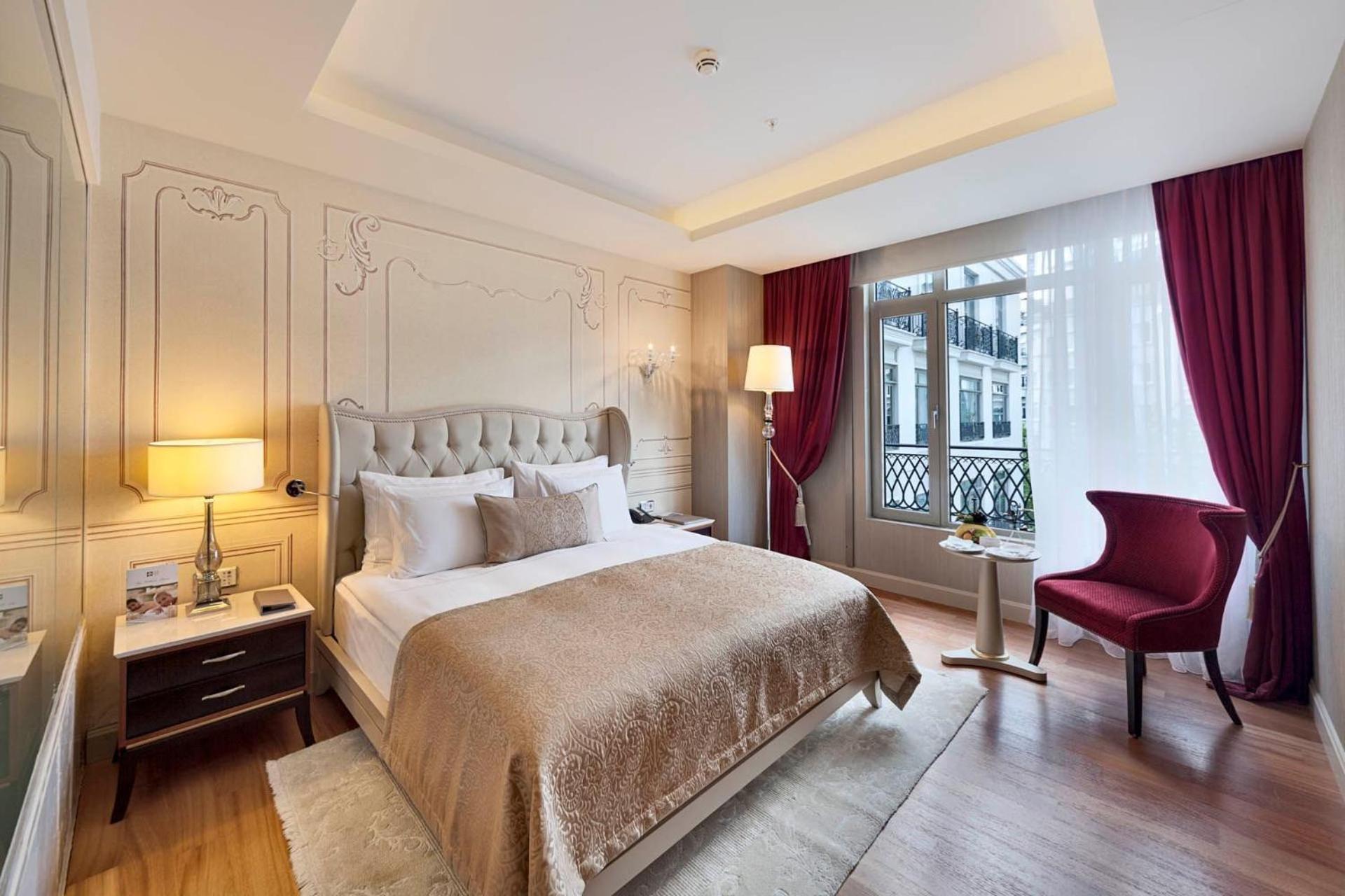 Cvk Park Bosphorus Hotel Istanbul Exterior photo The photo shows a well-decorated bedroom featuring a large bed with a beige and gold-colored bedspread. There are white pillows arranged neatly on the bed. Beside the bed, there are two nightstands with lamps. Across from the bed, there is a red acce
