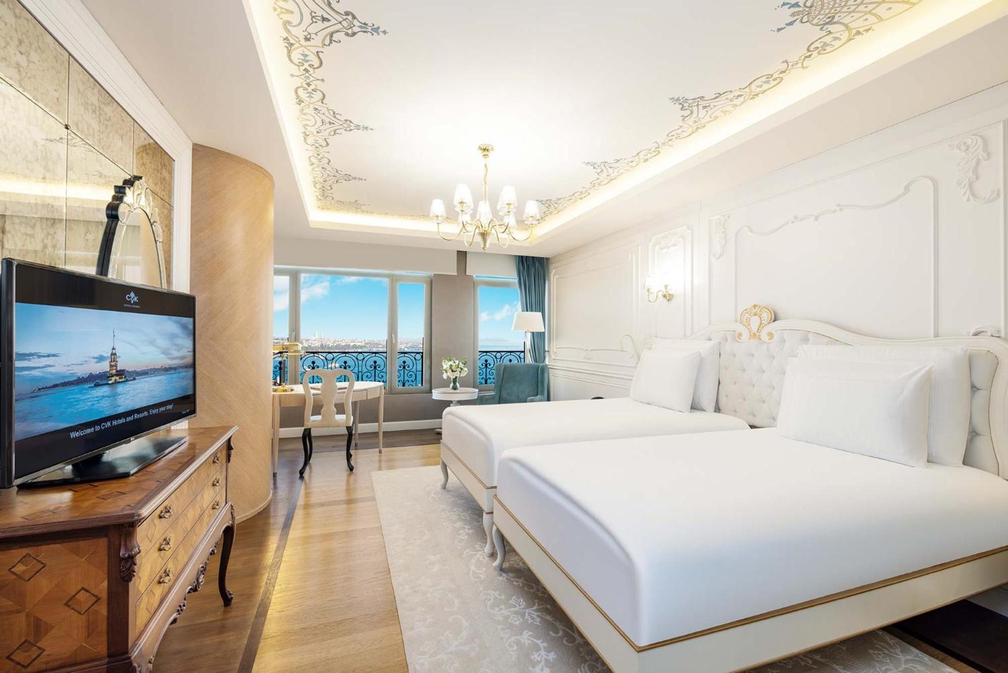 Cvk Park Bosphorus Hotel Istanbul Exterior photo The photo shows an elegantly designed hotel room featuring two twin beds with white bedding. The room has a luxurious feel with ornate ceiling details and soft lighting from a central chandelier. A wooden dresser with a television is positioned next 