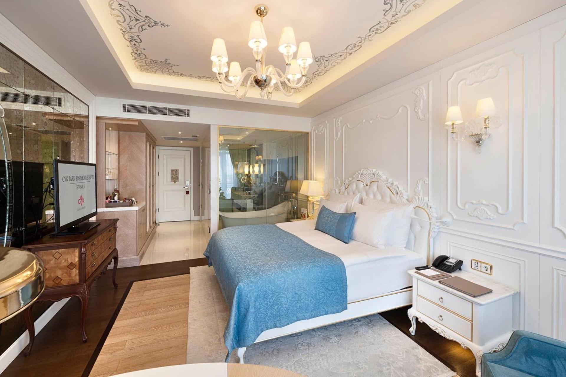 Cvk Park Bosphorus Hotel Istanbul Exterior photo The image shows a luxurious hotel room that features an elegant design. The room has a plush bed dressed in white linens and a light blue throw blanket. There is a decorative headboard and ornate molding on the walls. 

In the corner, a flat-screen t