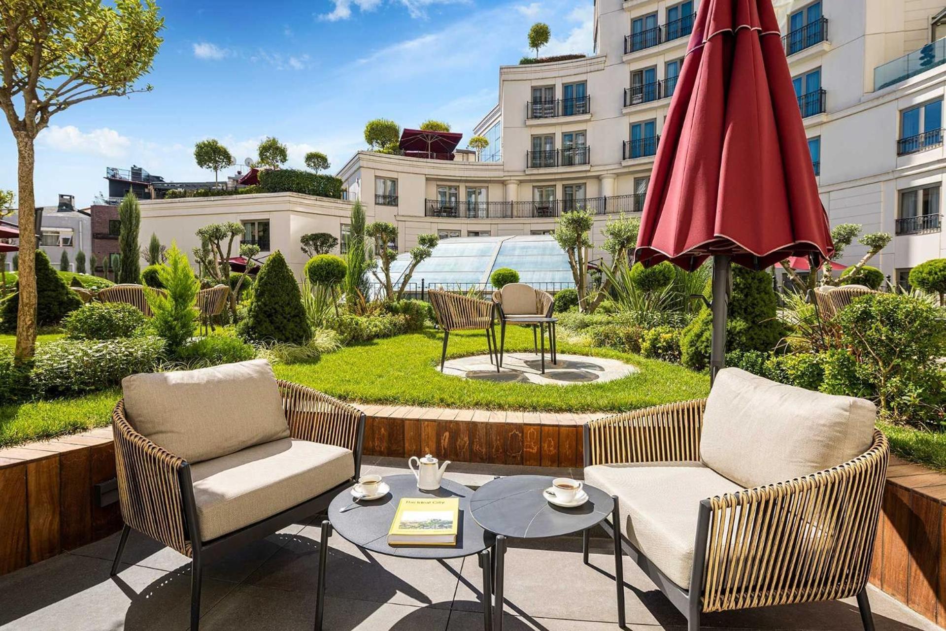 Cvk Park Bosphorus Hotel Istanbul Exterior photo The photo depicts a charming outdoor terrace area featuring two cozy chairs and a small round table. On the table, there are two cups, a teapot, and a magazine or book. In the background, there's a beautifully landscaped garden with various plants an